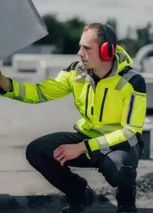 Arbeiter mit Warnschutzjacke und Gehörschutz, kniend auf Baustelle, Fokus Arbeitssicherheit