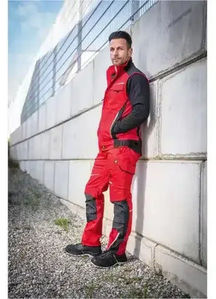 Mann in roter Arbeitskleidung mit schwarzem Einsatz, steht an Betonwand, Outdoor, Arbeitsschutz