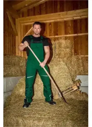 Mann in grüner Arbeitskleidung steht mit Heugabel in einer Scheune auf Strohballen