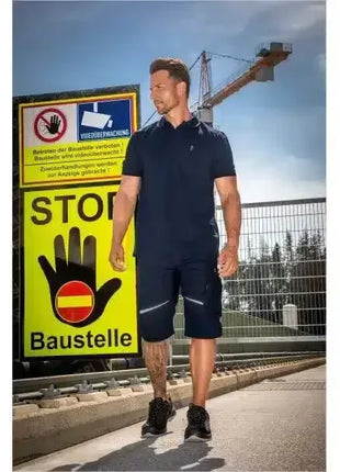 Mann in navyblauer Arbeitskleidung auf Baustelle vor STOP-Schild und Kran bei sonnigem Wetter