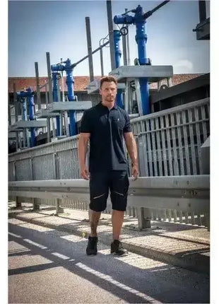Mann in Arbeitskleidung, schwarze Shorts und Polo, läuft auf Industriegelände mit Metallzäunen