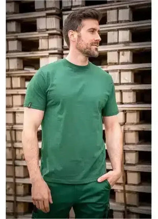 Mann in grünem T-Shirt und Arbeitskleidung steht vor gestapelten Paletten, Berufsbekleidung