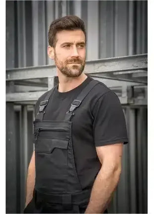 Mann mit Bart trägt schwarze Arbeitslatzhose und T-Shirt, industrieller Hintergrund