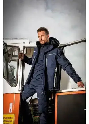 Mann in dunkler Arbeitsbekleidung mit reflektierenden Streifen steht in geöffneter Tür eines Fahrzeugs, Outdoor, Berufsbekleidung.