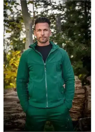 Mann im grünen Fleecejacke und Hose steht draußen vor Holzstapeln, Herbstwald im Hintergrund