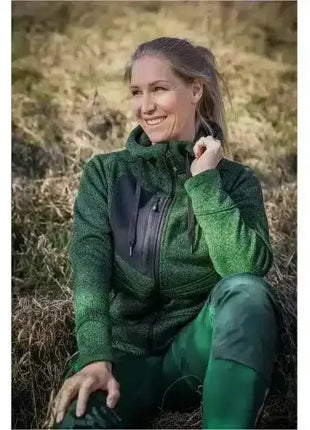 Frau in grüner Outdoor-Jacke sitzt lächelnd im Freien, natürliche Herbstumgebung, funktionale Kleidung