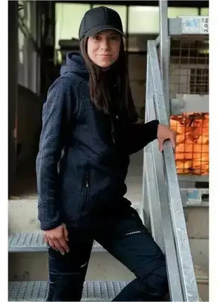 Frau in dunkelblauer Arbeitskleidung, Kapuzenjacke und Kappe auf Industrietreppe