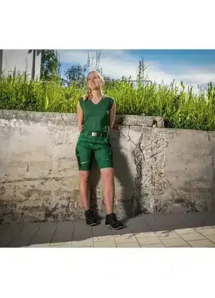 Frau in grüner medizinischer Arbeitskleidung steht draußen vor einer Betonmauer, Sonnenschein.