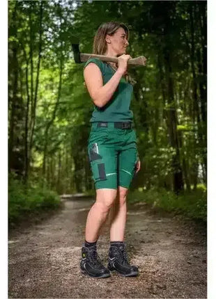 Frau in grüner Arbeitskleidung mit Axt auf Waldweg, Sicherheitsschuhe, Outdoor-Arbeitsbekleidung