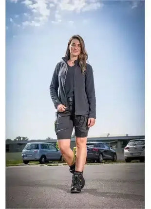 Frau in moderner Arbeitskleidung mit Softshelljacke und Shorts, draußen auf Parkplatz, professionelle Berufsmode.